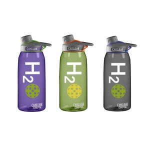 May include: Three CamelBak Chute water bottles in purple, green, and gray. Each bottle features the text "H2" in white, a pickleball graphic, and the CamelBak Chute logo. The lids have green, orange, and blue accents.