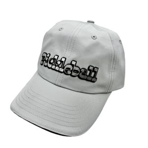 May include: A light grey baseball cap featuring the word "Pickleball" embroidered in black and silver. The cap has a curved peak and a button on the crown.