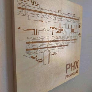 May include: A wooden sign with a laser-cut design of the Phoenix Sky Harbor International Airport (PHX) layout. The sign is light brown and features the airport code "PHX" and the city "Phoenix, AZ" in a darker brown.
