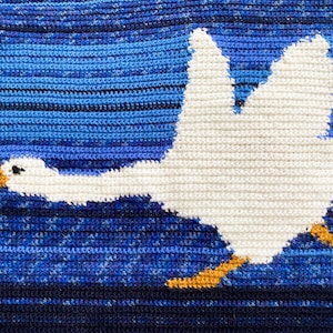 Könnte beinhalten: Eine weiße gehäkelte Gans fliegt über einem blau-schwarzen gestreiften Hintergrund. Die Gans hat gelbe Füße und einen gelben Schnabel.