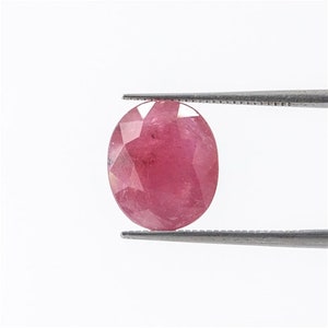May include: A pink oval-shaped gemstone with a faceted cut. The gemstone is held by a pair of tweezers.