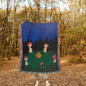 May include: A woven tapestry throw blanket with a dark blue background and a starry night sky. The blanket features a scene of four mushroom-shaped characters dancing around a campfire in a forest. The characters are white with red mushroom caps and are dancing in a circle around a small orange campfire. The forest is depicted in black silhouettes against a dark blue sky. The blanket has a green field with a fringe border.