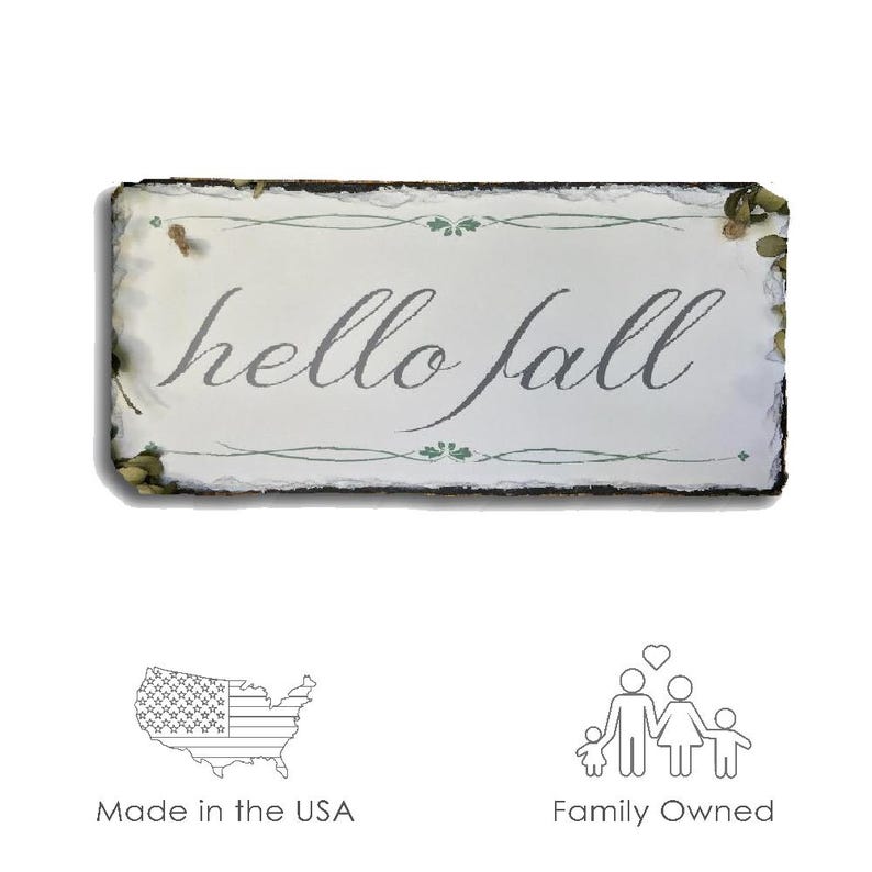 May include: A rustic wooden sign with a white background and the text "hello fall" in gray lettering. The sign has a distressed look and is decorated with green leaves and vines.