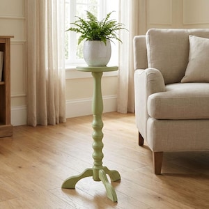 May include: A light green plant stand with a round top and a decorative base. A white ceramic pot with a green plant sits on top. The stand is approximately 30 inches tall.