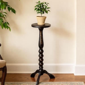 May include: A tall, dark brown wooden pedestal plant stand with a circular top and a central turned column featuring a series of rounded, stacked decorative elements. The stand rests on three curved legs and holds a small green plant in a beige pot.