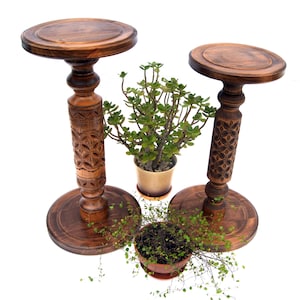 Set of two tall carved wooden plant pedestals in a dark finish. The pair includes one taller and one shorter stand with ornate turned columns and geometric carvings, shown holding potted ivy and fern plants on a rustic jute rug.