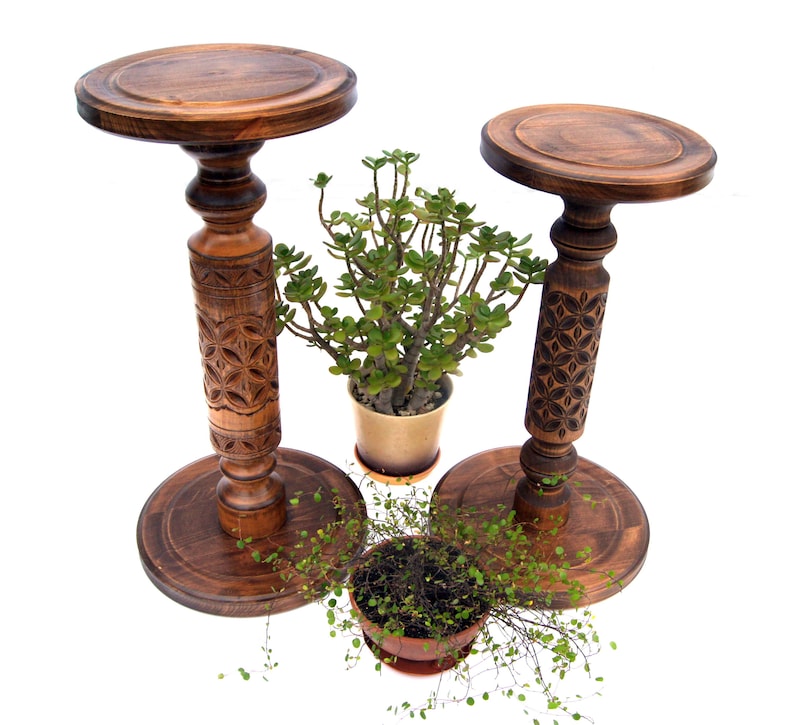 Set of two tall carved wooden plant pedestals in a dark finish. The pair includes one taller and one shorter stand with ornate turned columns and geometric carvings, shown holding potted ivy and fern plants on a rustic jute rug.