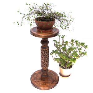 May include: A wooden plant stand with a carved design on the pedestal. The stand has a round top and a round base. Two potted plants are on the stand, one with green leaves and the other with small, round leaves.