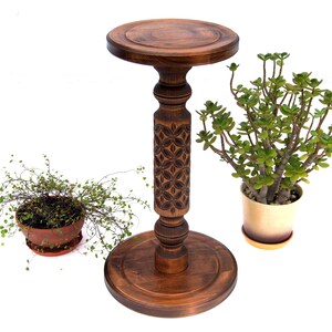 May include: A dark brown, wooden plant stand with a carved floral design on the central column. The stand has a round top and base. Two potted plants are on either side of the stand, one with small green leaves and the other with larger green leaves.