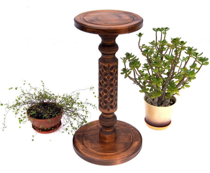 May include: A dark brown, wooden plant stand with a carved floral design on the central column. The stand has a round top and base. Two potted plants are on either side of the stand, one with small green leaves and the other with larger green leaves.