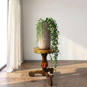 May include: A tall, beige ceramic vase with a textured surface, filled with a trailing green plant, sits atop a small, ornate wooden plant stand. The stand has a round top and a carved, three-legged base, showcasing a natural wood finish.