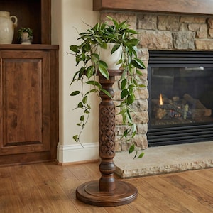 Op de afbeelding: Een hoge, donkerbruine houten plantenstandaard met een gesneden bloemmotief op de centrale kolom. Een witte keramische pot bevat een groene bladplant met ranken. De standaard staat voor een open haard.