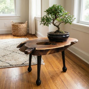 May include: A bonsai tree in a black pot rests on a live-edge wooden table. The table has dark, rounded legs and a natural wood grain finish. A woven basket and a rug are in the background.