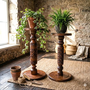 Rustic bohemian wooden pillar stands for indoor plants. A tiered set of two dark, ornately carved display pedestals styled in a sunlit cottagecore setting with stone walls, terracotta pots, and garden decor.