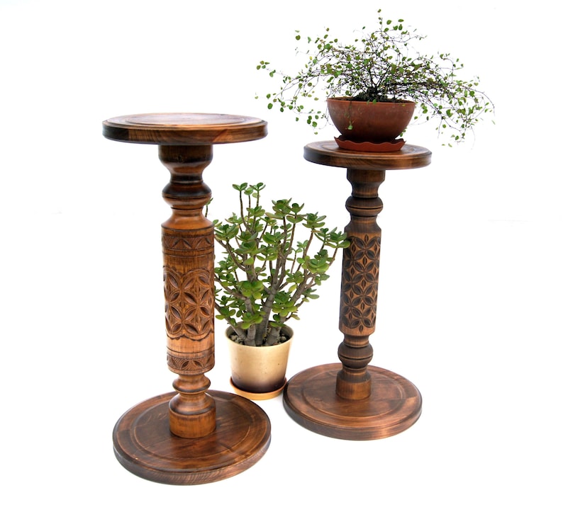 Set of two tall carved wooden plant pedestals in a dark finish. The pair includes one taller and one shorter stand with ornate turned columns and geometric carvings, shown holding potted ivy and fern plants on a rustic jute rug.