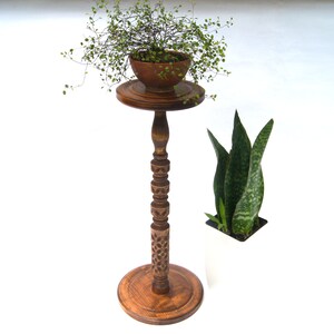 May include: A wooden plant stand with a carved design and a round top. A small potted plant sits on the top of the stand. A second plant is in a white pot on the floor.
