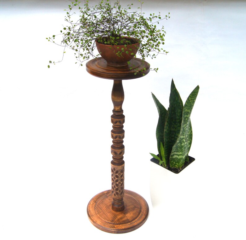May include: A wooden plant stand with a carved design and a round top. A small potted plant sits on the top of the stand. A second plant is in a white pot on the floor.