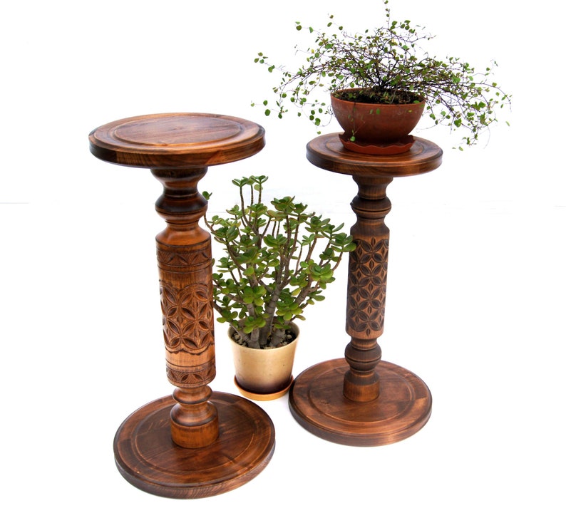 May include: Two wooden plant stands with intricate carvings. The stands are brown and have a round top. The stands are decorated with a floral pattern. The stands are on a white background.