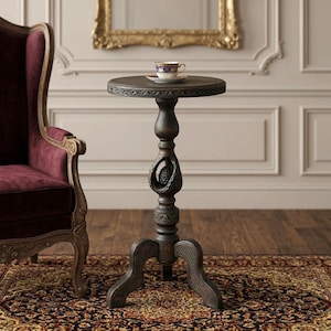 May include: A dark brown, antique-style side table with a round top and carved details. The table has a three-legged base and a decorative central support. A small teacup and saucer with gold trim sit on top. The table is approximately 24 inches tall.