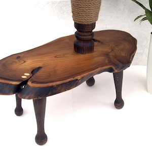 Op de afbeelding: Een kleine houten kruk met een natuurlijk blad met boomrand, zichtbare donkere houtnerf en een centrale barst gevuld met twee lichtgekleurde ronde inlays. De kruk staat op vier donkere, gedraaide houten poten met ronde voeten.