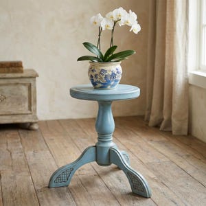 May include: A light blue wooden pedestal table with a round top and three curved legs, featuring a white ceramic planter decorated with a blue willow pattern and a white orchid plant with green leaves resting on the table surface.