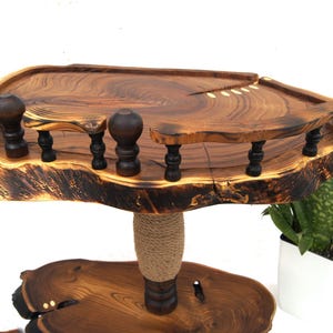 Op de afbeelding: Een houten standaard met twee niveaus, bestaande uit natuurlijke boomstam-schijven met een gepolijste afwerking. Het bovenste niveau heeft een donkerbruine decoratieve reling met gedraaide spijlen. Een centrale steunpilaar is omwikkeld met jutekoord.