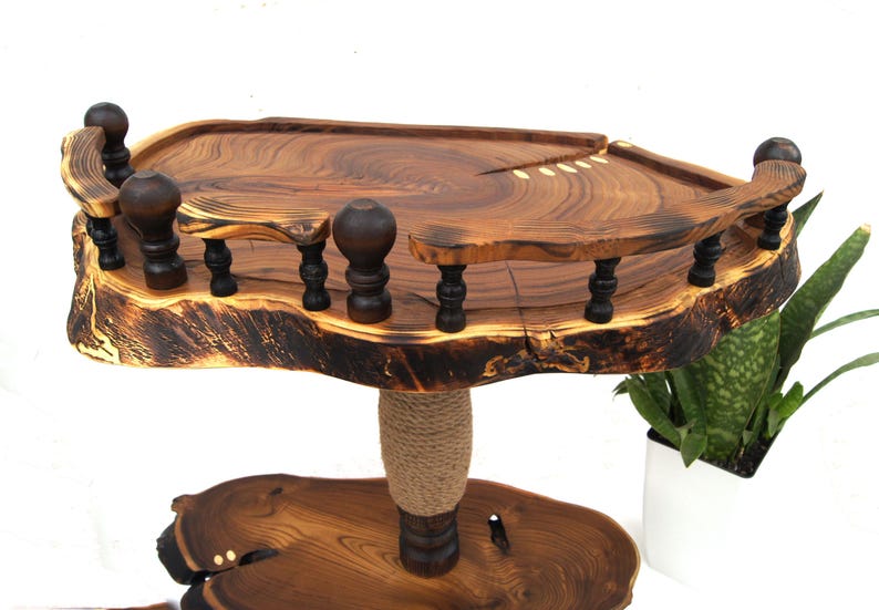 Op de afbeelding: Een houten standaard met twee niveaus, bestaande uit natuurlijke boomstam-schijven met een gepolijste afwerking. Het bovenste niveau heeft een donkerbruine decoratieve reling met gedraaide spijlen. Een centrale steunpilaar is omwikkeld met jutekoord.