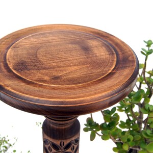 May include: A handcrafted, dark brown wooden pedestal with a round top and carved floral details on the base. The pedestal is designed to display items and is surrounded by green plants, creating a natural aesthetic.