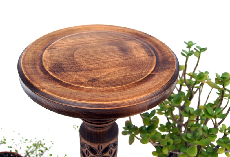 May include: A handcrafted, dark brown wooden pedestal with a round top and carved floral details on the base. The pedestal is designed to display items and is surrounded by green plants, creating a natural aesthetic.