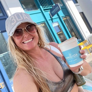 May include: A woman wearing a grey baseball cap with the number 30A on it, sunglasses, and a silver metallic sports bra, is holding a white to-go coffee cup with the logo "AMAVIDA COFFEE ROASTERS" printed on it.