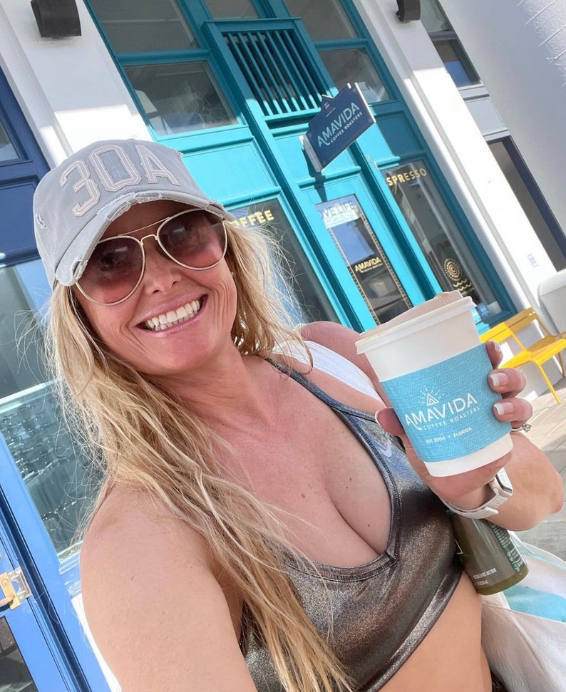 May include: A woman wearing a grey baseball cap with the number 30A on it, sunglasses, and a silver metallic sports bra, is holding a white to-go coffee cup with the logo "AMAVIDA COFFEE ROASTERS" printed on it.