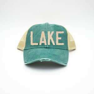 May include: A green and beige trucker hat with the word "LAKE" in beige lettering on the front.