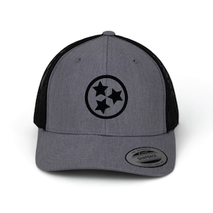 May include: A gray and black trucker hat with a black circle and three stars in the center. The hat has a black mesh back and a gray front. The text "The Authentic Snapback" is on a white tag attached to the hat.