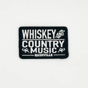 May include: A black rectangular patch with white embroidered text that reads "Whiskey and Country Music Nashville". The patch has decorative flourishes and a white border. The patch is likely for a jacket or bag.