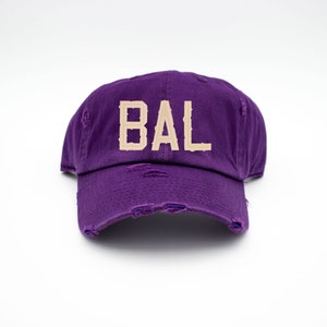 May include: Purple distressed baseball cap with the word "BAL" in beige embroidered on the front.
