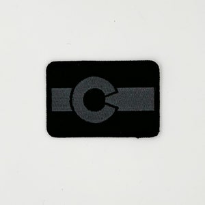 May include: Black and gray embroidered patch with a stylized letter "C" in the center. The letter is surrounded by a gray band.