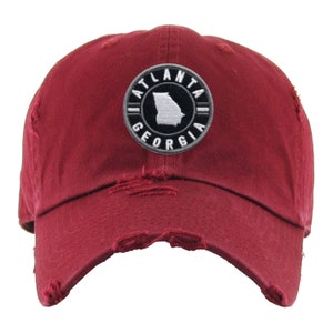 May include: A maroon distressed baseball cap with a black and white embroidered patch that says "Atlanta Georgia" and features an outline of the state of Georgia.