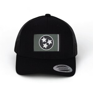 May include: Black trucker hat with a green and white Tennessee state flag patch. The hat has a black mesh back and a black snap closure. The text "The Authentic Snapback" is printed on the back of the hat.