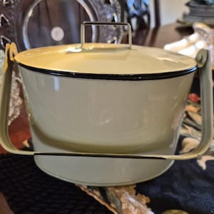 May include: A vintage, cream-colored enamelware bucket with a lid and a black trim. The bucket has a metal handle and a matching handle on the lid. The bucket is sitting on a patterned surface.