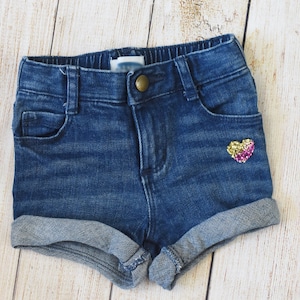 May include: Blue denim shorts with a rolled hem. The shorts have a button closure and a small pocket on the front. A sequined heart in gold and pink is on the right pocket. The shorts are designed for a child.