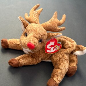 Puede incluir: Un peluche de reno con pelaje marrón, una nariz roja y grandes cuernos. El juguete tiene una etiqueta roja en forma de corazón que dice "2000". El reno está tumbado con las patas estiradas.