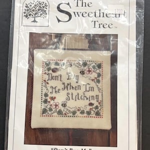 Może przedstawiać: Zestaw do haftu krzyżykowego w przezroczystym plastikowym opakowaniu. Zestaw zawiera haftowany wzór z napisem "Don't Bug Me When I'm Stitching" otoczony motywami kwiatowymi i owadami. Opakowanie zawiera markę "The Sweetheart Tree."
