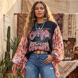 May include: Black graphic tee with "Cowboys" text and a cowboy illustration. The sleeves are a light pink floral pattern. The model is wearing blue jeans and a woven belt. Turquoise jewelry and rings complete the look.