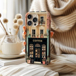 May include: A phone case featuring a detailed illustration of a coffee shop with the word "COFFEE" above the entrance. The design includes windows, potted plants, and a charming architectural style, set against a soft, neutral background.