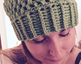 May include: A close-up of a woman wearing a green crocheted beanie hat. She is wearing a white shirt and a silver necklace with a pendant.