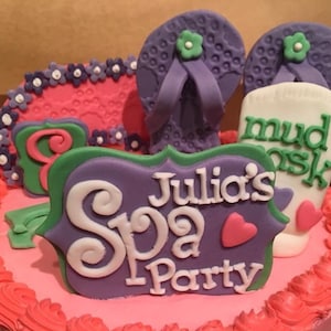 May include: A pink and white cake decorated with purple and green fondant. The cake is decorated with a sign that says "Julia's Spa Party", flip flops, and a tube of mud mask.