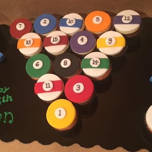 May include: A black and white cake decorated with cupcakes resembling billiard balls. The cupcakes are arranged in a triangle shape, with the numbers 1 through 15 on each cupcake. The cake is decorated with the words "happy 35th Ryan".