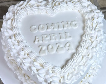 May include: White heart-shaped cake decorated with white frosting and the words "Coming April 2024" in white frosting.