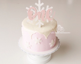 May include: A small, round, pink cake with white icing and snowflake decorations. A white and pink snowflake cake topper spells out "one". The cake is on a white, scalloped-edge cake stand. Likely for a first birthday celebration.