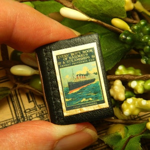 Peut inclure: Petit livre noir intitulé "Every Boy's Book of Railways & Steamships" avec une illustration colorée d'un navire. Le livre est tenu par des doigts. Le fond est orné de verdure et d'éléments décoratifs.